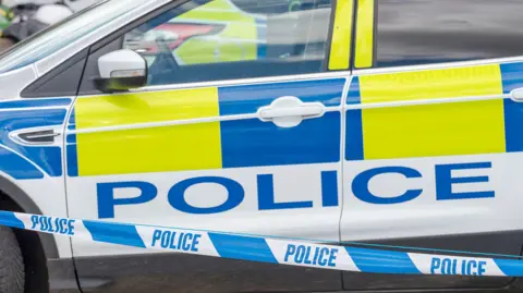 Getty Images The side view of a police car with blue and yellow markings. A blue and white police cordon crosses in front of it. 