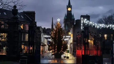 Calum Taylor A lit up Christmas tree sits in the middle of a town centre. Other Christmas lights are also hanging to the side.