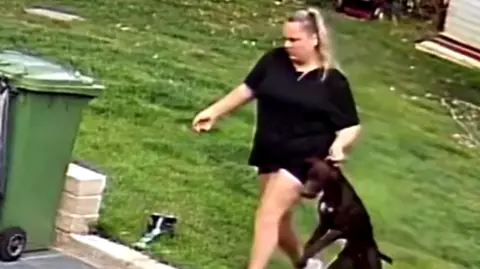 A woman can be seen dragging a black dog onto a patio area with a lawn behind. There is a green wheelie bin on the left hand side of the image. The woman is wearing a black top and has blonde hair.
