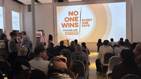 Kate Bradbrook/BBC People are sitting on rows of chairs in a room looking towards the front where a projector is showing an image that says 'no one wins' on it.