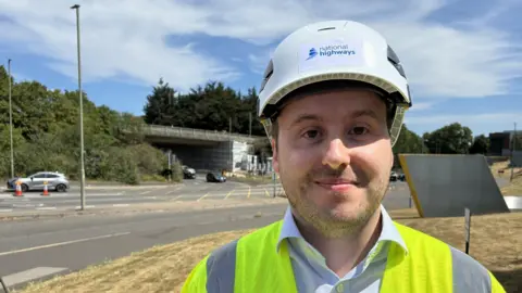 A man wearing a blue shirt, yellow high-vis vest and white hard hat with the national Highways logo on the front. He is smiling at the camera. Over his right shoulder is a roundabout