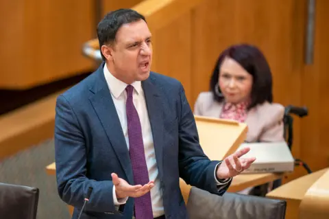 PA Media Anas Sarwar stands up in the debating chamber in the Scottish parliament. he wears a dark grey suit and speaks and gestures with his hands