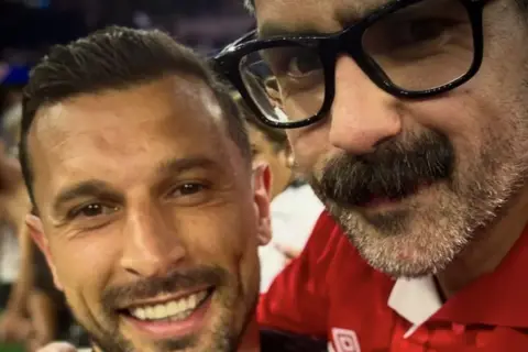Gav Murphy Kosta smiles with Gav's arm around him, Gav wearing a Wales jersey. Gav has a moustache and short trimmed beard and Kosta has short trimmed facial hair