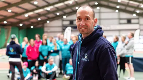 Gergana Marinova Tim Howe standing in front of a group of people in a sports hall. He smiles at the camera and wears a dark-blue hoodie which has The Longevity Games logo on it.
