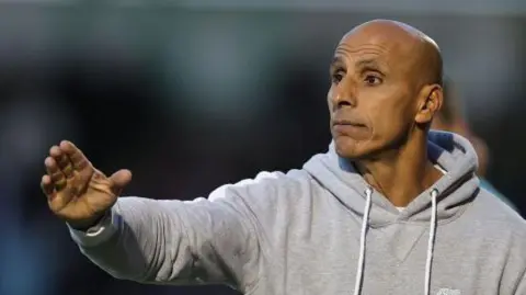 Dino Maamria pointing with his right hand, wearing a grey hoodie 