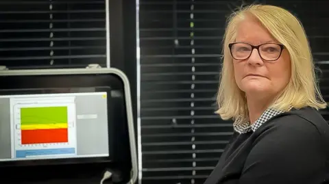 Jill Dalgleish has blonde shoulder length hair, dark rimmed glassed and it wearing a herring bone patter top under a dark jumper. she is standing in a doctor's office next to a screen showing bone density 