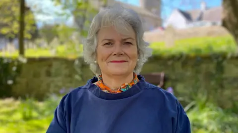 Sue Aldwell, infront of greenery, wearing a navy top and an orange patterened shirt under it. She has medium length grey hair.