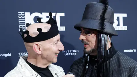 Getty Images A photo of Stephen Jones and Boy George from the shoulders up. Jones stands on the left, he has a bald head and is wearing a black crown made from a felt-style fabric. His jacket has a newspaper print and he wears a black top underneath. Boy George is wearing a pinstripe jacket and a black bucket-style hat with tassles. He has facial hair and gems around his eyes. 