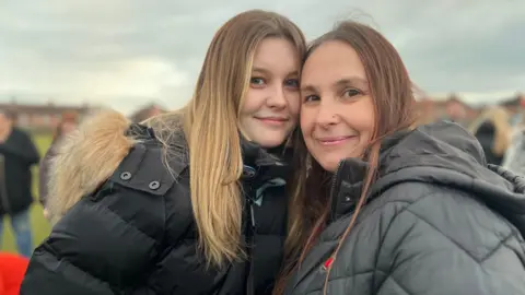 A mum and her young adult daughter both smile at the camera. They are both wearing coats as they stand on a field at a community centre, with a crowd behind them. 