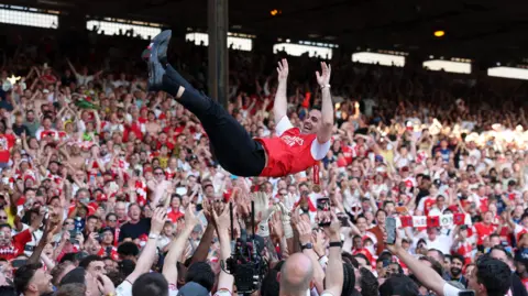 Getty Images Mikel Arteta is thrown into the air by his players in front of a packed crowd of Arsenal supporters, arms outstretched mid-celebration.