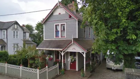 Google A grey-green painted house in an American clapboard style with a porch running around it and lots of trees in the front garden, bounded by a matching coloured board fence