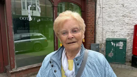 A woman in a blue coat and yellow jumper looks at the camera outside a glass fronted building.