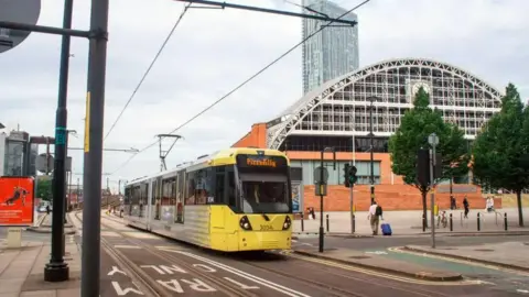 A yellow tram with Piccadilly on the front passes Manchester Exhibition Centre heading towards St Peter's Square tram stop