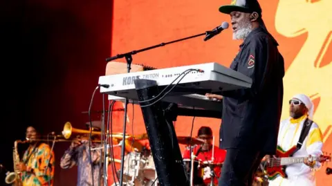 Steel Pulse perform on stage at the 2024 Glastonbury Festival. In the foreground the keyboardist dominates the shot, but behind him can be seen a guitarist, drummer, a trombonist and a saxohphonist.