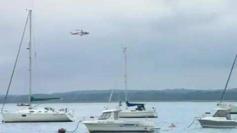 Sailing boats in the sea, with a coastguard helicopter overhead.