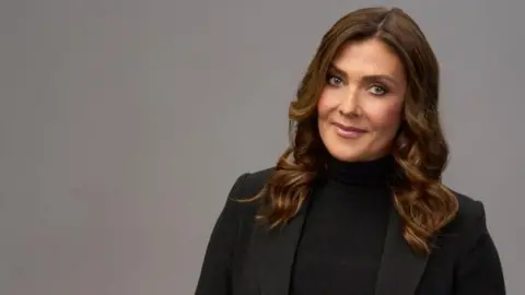 Kym Marsh, who is wearing a black blazer and black polo neck jumper, is standing in front of plain grey background for this posed photograph and is smiling. 