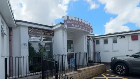 The entrance of a school. There is fencing around a walkway to the front door. The front end of a grey car can be seen on the right. Two signs reading "Moat Farm Junior School" are on the front of the building.