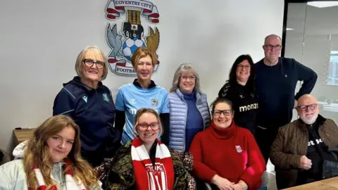 Coventry City Disabled Supporters Association A group of six women and two men all of different ages are arranged in a row of two, the front row is sitting down and the back row are standing behind them. In the background on a white wall is a Coventry city logo