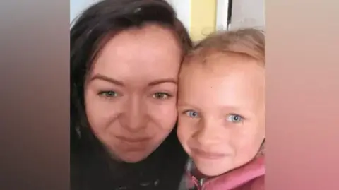 A woman with long dark hair and brown eyes is cuddled up next to a young girl with blonde hair and blue eyes wearing a pink top.