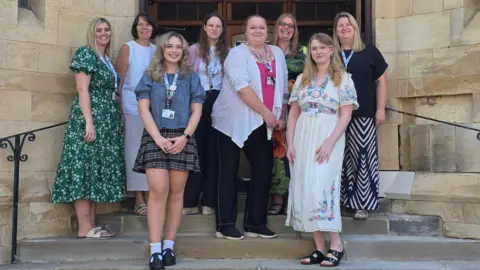 A group of women, all standing outside a stone building, on its steps, smiling and looking straight at the camera. They are all by a large wooden door, and mostly have lanyards on round their necks.