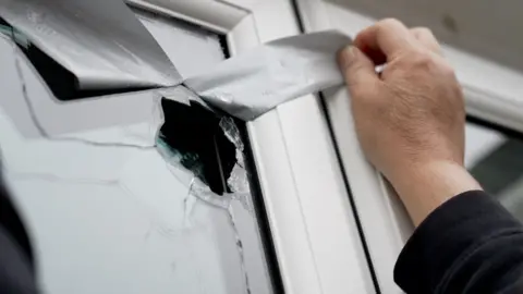 Ben Moore/BBC A man is pulling back the tape on a cracked window to show the damage caused by a projectile fired by a catapult.