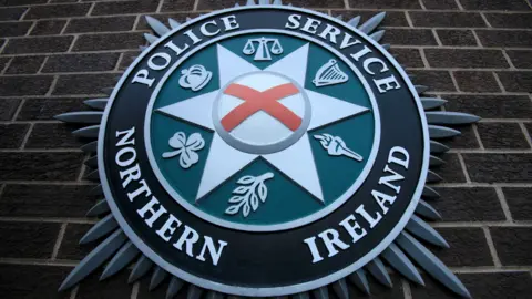 The PSNI badge features the St. Patrick's saltire, and six symbols The Scales of Justice, a harp, a torch, a laurel leaf, a shamrock, and a crown. It's attached to a brick wall.