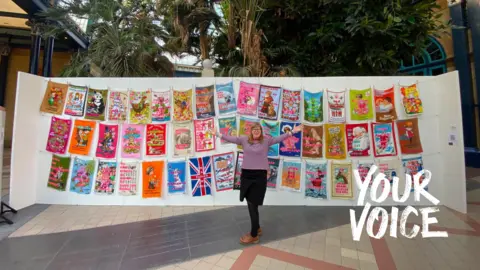 Large display of colourful tea towel artworks hung on lines, with one person standing in front of the installation.