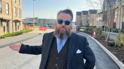 Shariqua Ahmed/BBC Dan Lentell stands on a road being built on a new housing development. He is wearing dark glasses with blue frames and has a greyish-brown beard. He is wearing a white shirt, a grey blazer with a white handkerchief in the breast pocket and a blue tie. He is holding out his right hand as if expressing exasperation.