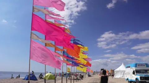 Colourful flags of pink, yellow, orange and blue have been placed in a line across the seafront of the beach and are flying in the wind. There are people on the beach sitting down with chairs and tents and people walking. 