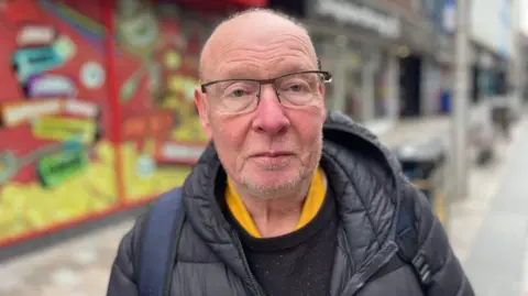 Tony Cusack wears glasses. He has a coat and jumper on. A backpack is over his back. A store is behind him.