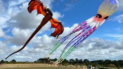 A red dragon and white octopus kits flying over a large flat open field with trees on the horizon.