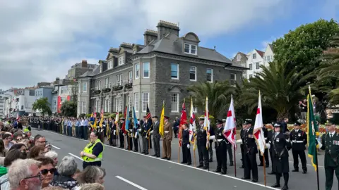 BBC Crowds behind a barrier on the left look across at a military and civilian parade with standard bearers carrying the flags of various groups, organisations and military units in the foreground with other groups further back. Behind the parade are building along St Peter Port seafront.