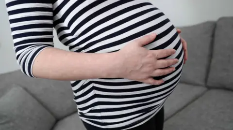 The image shows a close-up view of a pregnant person's bump, with both hands resting on the stomach while wearing a navy and white striped top. In the background there is a grey sofa, and the photograph appears to have been taken indoors using soft natural light, with the focus centred on the pregnancy bump rather than the person's face, which is not visible.
