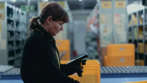 Conic Film We see the right side of a female warehouse worker who is holding and looking attentively down on a scanner. She's wearing a thick black jumper and a yellow lanyard, her hair is tied up. In the background we can see a conveyer belt and yellow warehouse boxes.