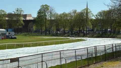 Grey covers on the racecourse, going from the bottom left to about the middle right of the photo. Trees are in the background. 