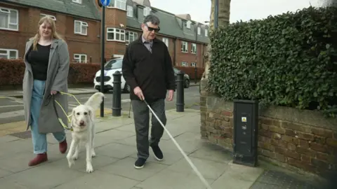 Jessica is walking along holding her guide dog beside Andrew who is using a white stick to help navigate the pavement