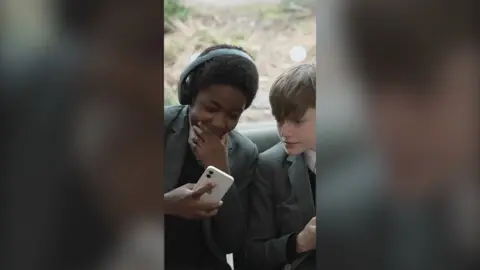 Two boys looking at a mobile phone. They are wearing grey school uniform. One boy is wearing headphones
