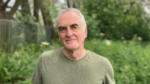 BBC Ash Smith looking at the camera - he's got white hair, black eyebrows and is wearing a green T-shirt. He's white.