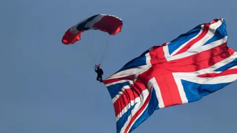 An action shot of the silhouette of a parachuter in freefall to the ground. The parachute is red, white and black and a large union jack flag is attached to them. The sky is blue in the background.