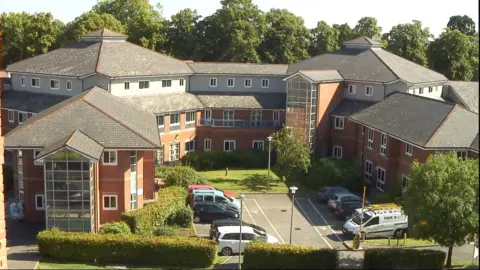 An aerial view of a large brick built hospital. There are cars parked in the car park at the front and the area is surrounded by trees with green leaves. 