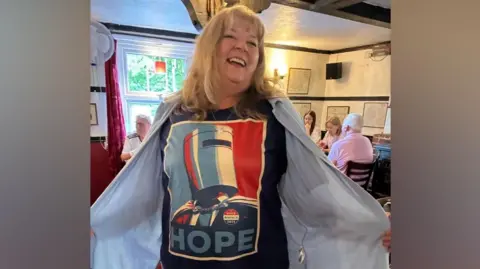 Una mujer muestra con orgullo su camiseta de Count Binface HOPE mientras está en un pub. Sonríe y mira a la cámara.