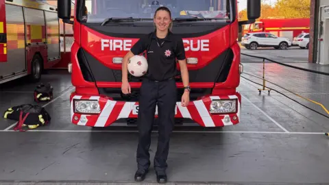 Bow Jackson stands in front of a red fire engine wearing Devon and Somerset Fire and Rescue branded clothing. She has a football with multiple Exeter City badges on it under her right arm.