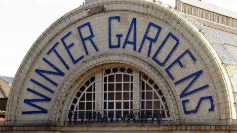 Semi circular tiled sign saying Winter Gardens in blue on cream on the outside of the building