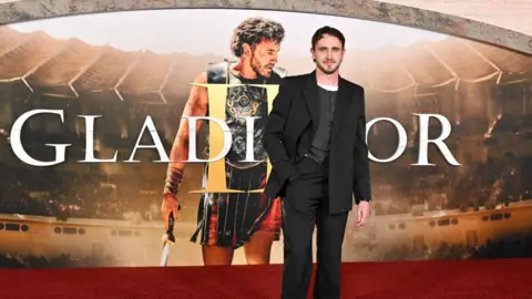 Getty Images Paul Mescal standing in front of a poster which has a photo of him in his gladiator costume and the words "Gladiator II" overlaid on top of it