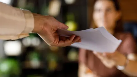 An outstretched hand holding white papers offering them to another outstretched hand. The background is blurred.