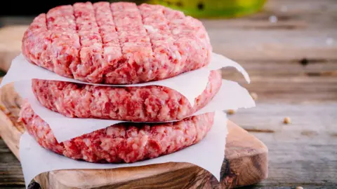 Three beef burgers separated by paper on top of a wooden chopping board and an wooden table