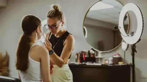 Getty Images A makeup artist with blonde hair tied in a high bun applies make up with a brush to the lips of a girl who can be seen from behind sitting in a chair in front of a mirror. A ring light is behind the make up artist and make up brushes and products are out of focus on a chest of drawers behind,