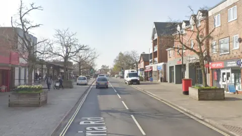 Google A screenshot from Google street view showing a road with wide pavements on either side. On the pavements are planters full of daffodils with trees in the middle.