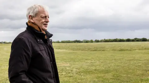 Dave Davies/SMGWA Nick is standing outside of the aerodrome.
He is wearing a dark jacket.
The weather is overcast.
The background is open green space.