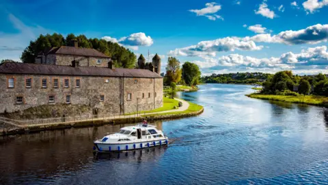 Getty Images A couple steer their Cruise Boat past Enniskillen Castle on Lough Erne Northern Ireland. A few people walk on the river's edge.